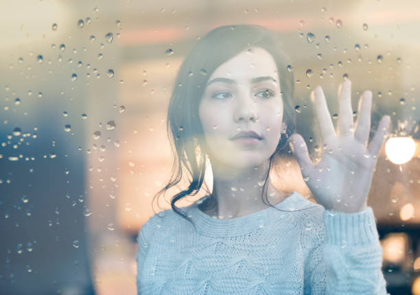 Sad young cute woman looking through window on rainy day