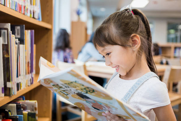 Happy little girl reads book in school library Adorable Hispanic schoolgirl smiles while delight as she reads a picture book while standing in the school library. reading stock pictures, royalty-free photos & images