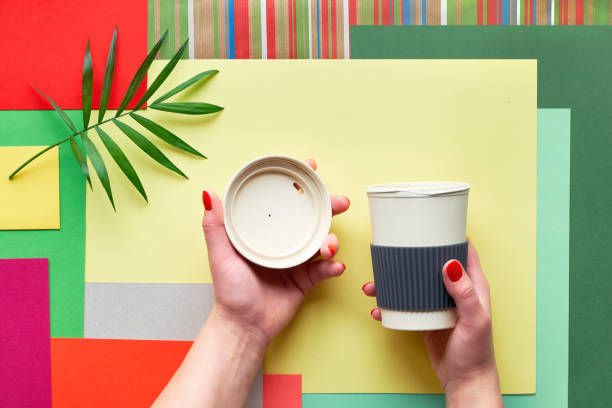 istock Bamboo coffee cup (keep cup or travel mug) with lid in female hands on abstract geometric multicolor rectangle paper background with palm leaf. Creative flat lay with zero waste biodegradable cup. 1193590438