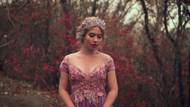 Portrait of a fantasy blonde hair cute princess. Woman in royal luxurious long purple dress with bare shoulders. Glamorous design fashion model. Silver crown. Elven queen in a fabulous autumn forest.