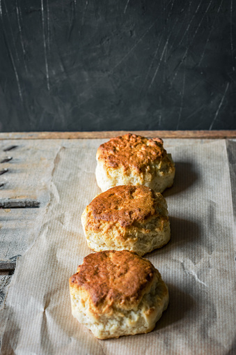 Fresh homemade English scones on parchment paper on aged wood table dark wall background. Traditional British pastry. Rustic kitchen interior
