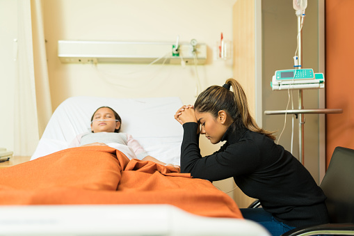 Tensed mother sitting by unconscious daughter lying on bed at hospital