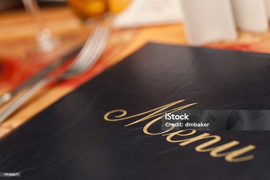 Menu on a restaurant table next to knife and fork A menu resting on a restaurant table, emphasis placed on the golden embroidery lettering against the black with the image's focus.  Silverware, napkins and glasses sit in the background as blurry shapes at the menu's edge, all resting on a light brown table. Menu Stock Photo