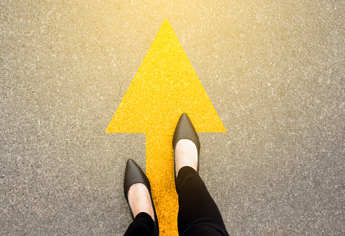 Feet and arrows on road background in starting line beginning idea. Top view. Business woman in black shoes on pathway with yellow direction arrow symbol. Moving forward, new start and success.