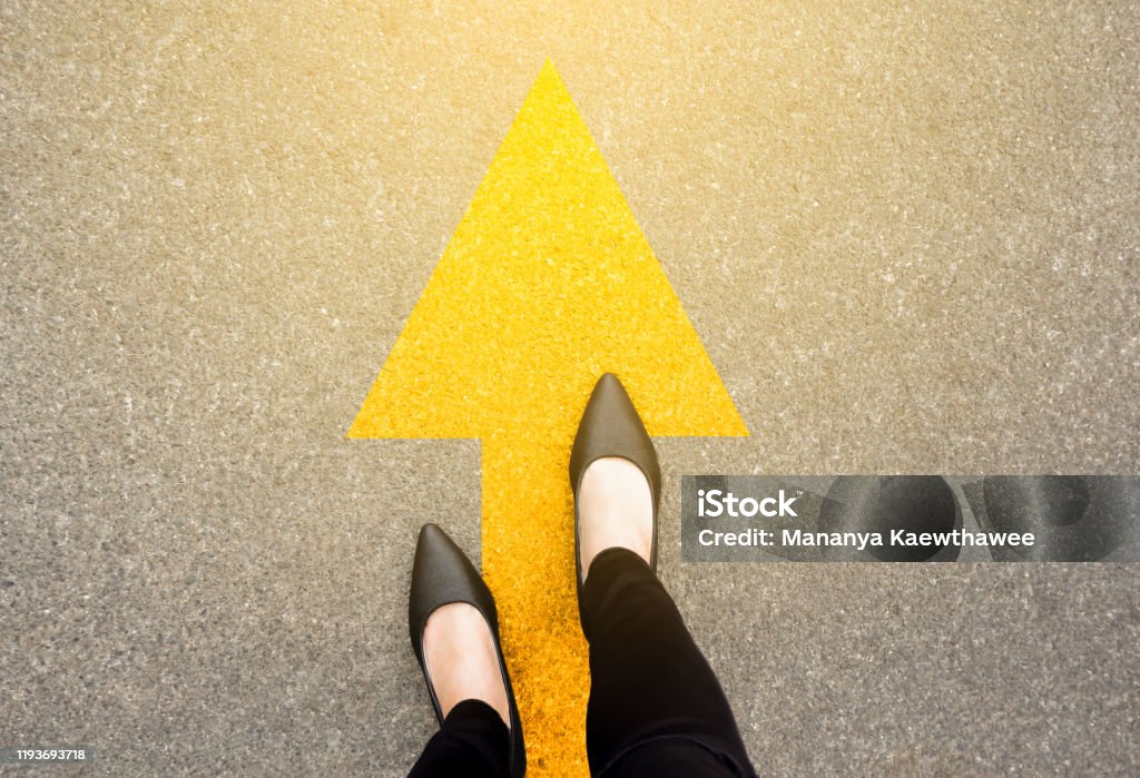 Feet and arrows on road background in starting line beginning idea. Top view. Business woman in black shoes on pathway with yellow direction arrow symbol. Moving forward, new start and success. Footpath Stock Photo