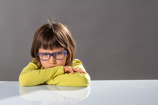 unhappy mad child with dirty look behind eyeglasses and arms folded on table expressing an irritated pouting no