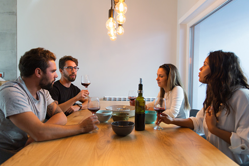 Group of friends arguing over glass of wine in dining room. Young men and women in casual meeting indoors. Emotional talk concept