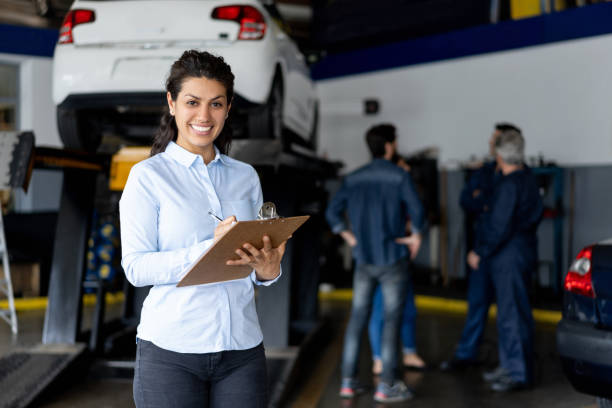 Friendly sales assistant at a car workshop holding a list on clipboard smiling at camera Friendly sales assistant at a car workshop holding a list on clipboard smiling at camera  - Incidental people at background auto mechanic photos stock pictures, royalty-free photos & images