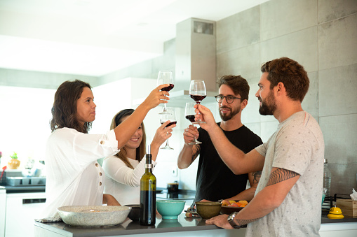 Happy positive friends celebrating event and toasting wine in kitchen. Young men and women in casual meeting indoors. Drinking wine at home concept