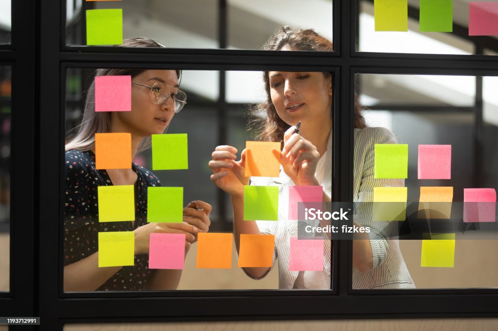 Two multiracial female partners planning business project. Two multiracial female partners planning business project, writing ideas on sticky notes. Mixed race colleagues checking working progress, adding tasks on glass wall, using kanban scrum methodology. Organization Stock Photo