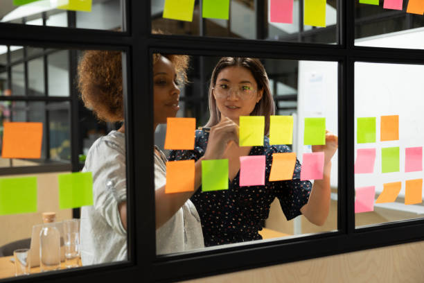 African american young female scrum master working with vietnamese colleague. Concentrated african american young female scrum master working together with smart vietnamese businesswoman colleague, putting notes on sticky paper at window glass agile kanban board at office. task stock pictures, royalty-free photos & images