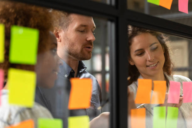 Close up headshot smiling diverse colleagues working near kanban board. Close up head shot smiling female african american and caucasian colleagues showing working progress to confident male team leader on kanban agile glass window board with colorful paper stickers. scrum stock pictures, royalty-free photos & images