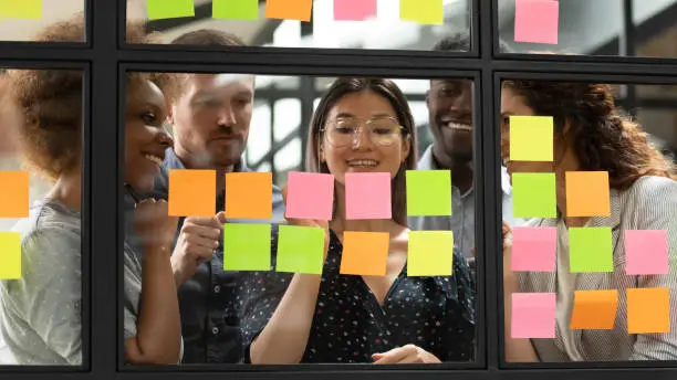 Mixed race interested young managers group gathering near smiling vietnamese team leader, writing startup project details or tasks on colorful sticky notes, using kanban scrum methodology at office.