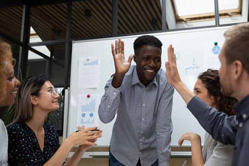 Happy african american team leader raising hand, giving high five to caucasian partner, investor or teammate at business people meeting in office. Mixed race young employees discussing project ideas.