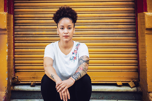 Portrait of woman sitting against closed yellow shutter. Confident female is having tattoo on hands. She is in fashionable casuals.