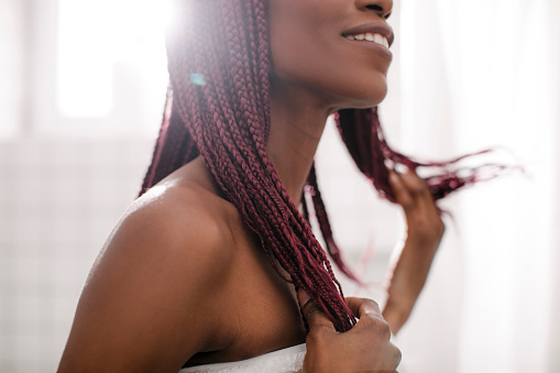 Beautiful smiling African woman standing at bathroom and holding her braided hair.