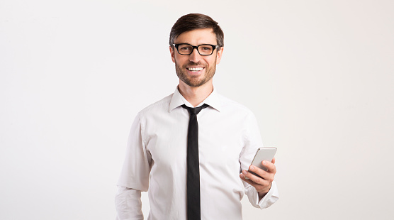Business App. Smiling Man Using Mobile Phone Looking At Camera Standing Over White Studio Background. Panorama, Empty Space