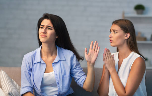 Girl Asking Indifferent Friend For Favor Sitting On Sofa Indoor Miserable Girl Asking Indifferent Friend For A Favor Begging Holding Hands In Prayer Gesture Sitting On Sofa Indoor. Selective Focus begging social issue photos stock pictures, royalty-free photos & images