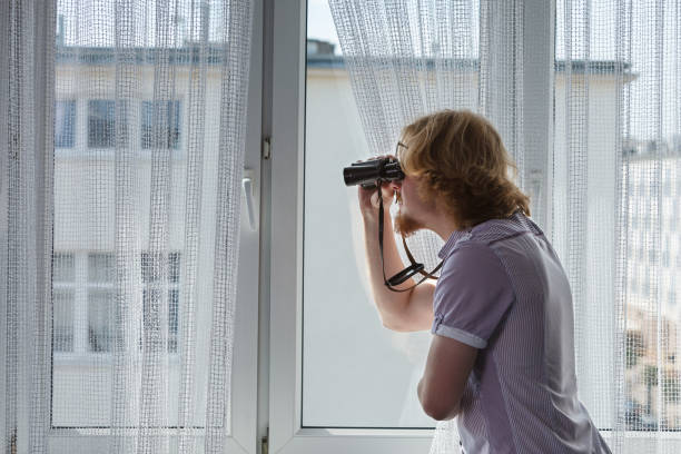Young adult man spying at his neighbors, guy using binoculars looking through window.