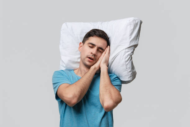 istock Tired brunet man in a blue tee resting on a white pillow isolated over grey background. 1193735740