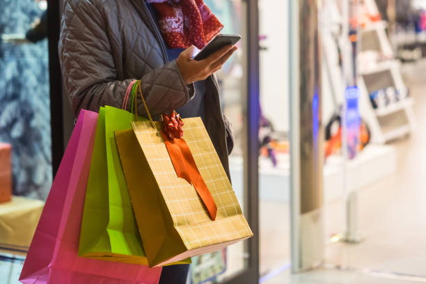 One people alone. A senior woman enjoys evening shopping while taking advantage of offers and discounts. On his arm a lot of shopping bags while she looks at her cell phone One people alone. A senior woman enjoys evening shopping while taking advantage of offers and discounts. On his arm a lot of shopping bags while she looks at her cell phone window shopping at night stock pictures, royalty-free photos & images