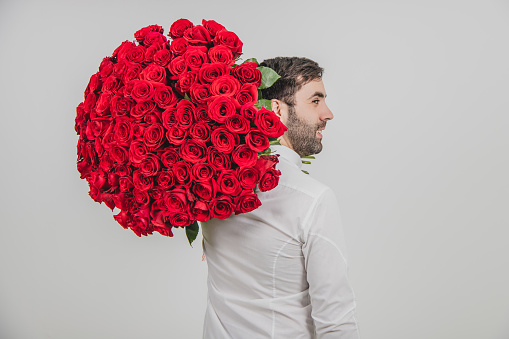 Man ready to make proposal concept. Portrait of confident man in formal clothes, standing back to the camera, carring a bouquet of red roses on his shoulder.