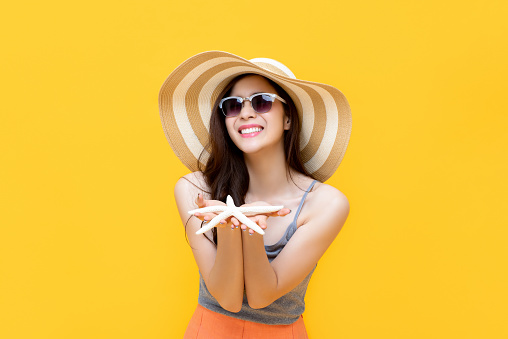 Beautiful young Asian woman in summer casual clothes smiling and holding starfish in hands isolated on yellow background