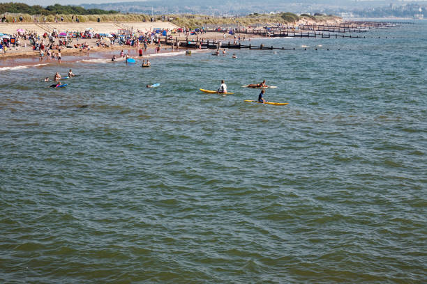 Looking across the water to the crowded beach with people in the sea.  Erosion prevention groynes in shot.