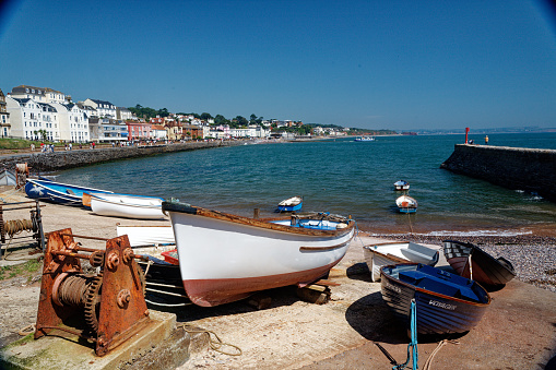 Colourful rowing boats on shore with small winch to their bow. View across blue water to town houses and seashore. Sunny day blue sky calm idyllic day.