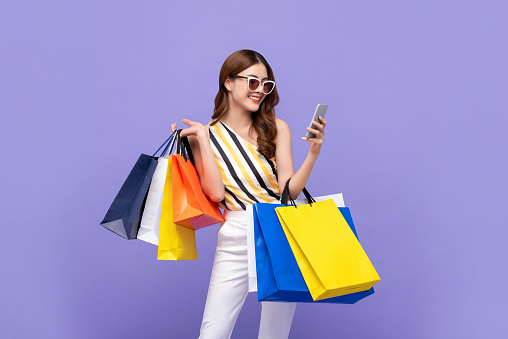 Trendy beautiful young Asian woman carrying colorful bags shopping online with mobile phone isolated on purple background
