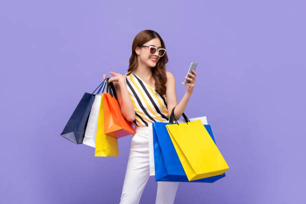 Beautiful Asian woman carrying colorful bags shopping online with mobile phone Trendy beautiful young Asian woman carrying colorful bags shopping online with mobile phone isolated on purple background shopping bag stock pictures, royalty-free photos & images