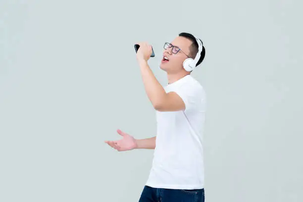 Young Asian man wearing headphones listening to music from mobile phone and singing isolated on light gray background