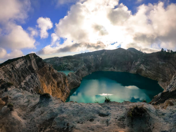 View on the Kelimutu volcanic crater lakes in Flores, Indonesia. Lakes are shining with many shades of turquoise and blue. Sun shines through clouds. Barren and sharp slopes of the volcanic crater