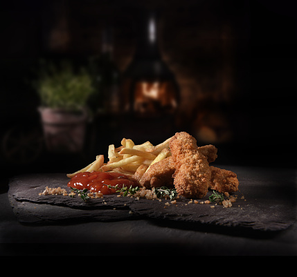 Succulent Chicken Nuggets, or guijons, covered with breadcrumbs, American style French fries and tomato ketchup and thyme herbs shot against a rustic background with generous accommodation for copy space.