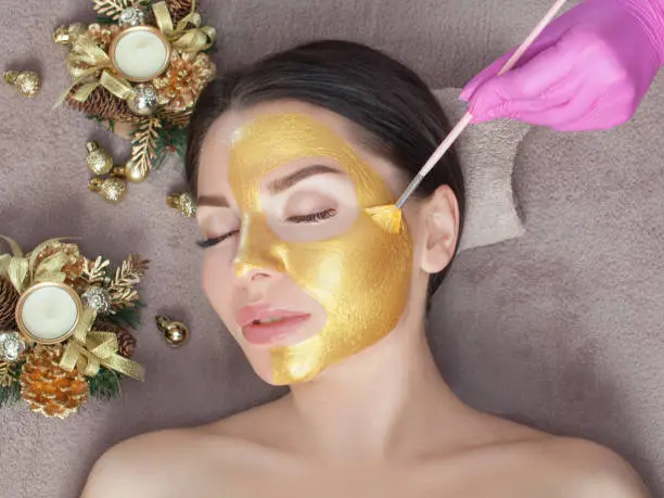 Beautician makes a golden mask to rejuvenate the skin to beautiful woman. Next to her are Christmas decorations.New Year's and Cosmetology concept.
