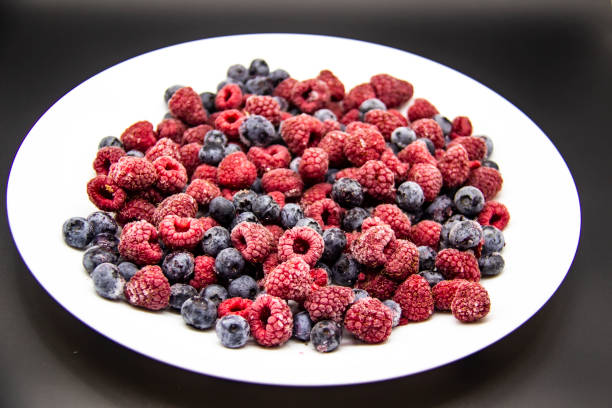 A Berry mix  from frozen raspberries and blueberries on the white plate. A Frozen Berries in black background.  A sweet background with frozen raspberries and blueberries