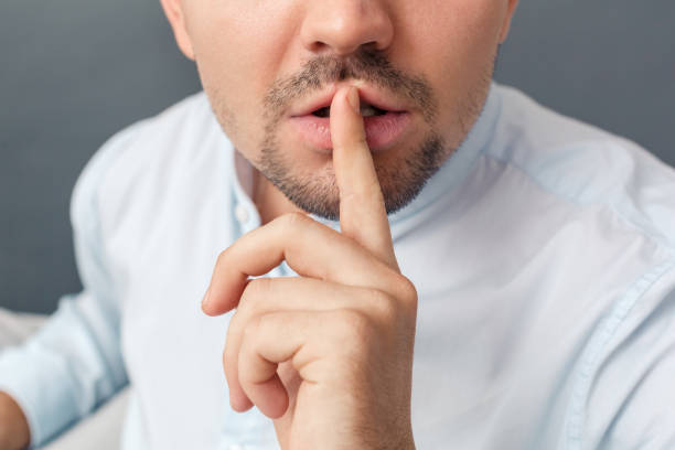 istock Be Quiet. Young man studio sitting on sofa isolated on grey showing hand gesture close-up 1193802590