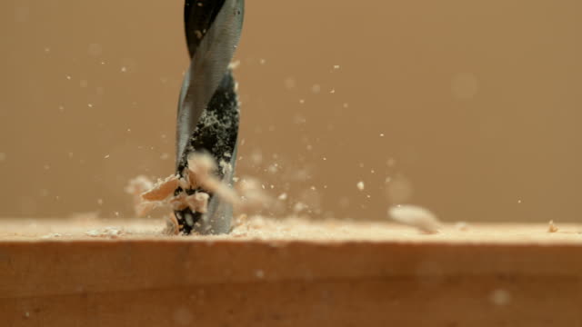 MACRO: Wood chips fly off a plank as handyman drills holes into the workpiece.