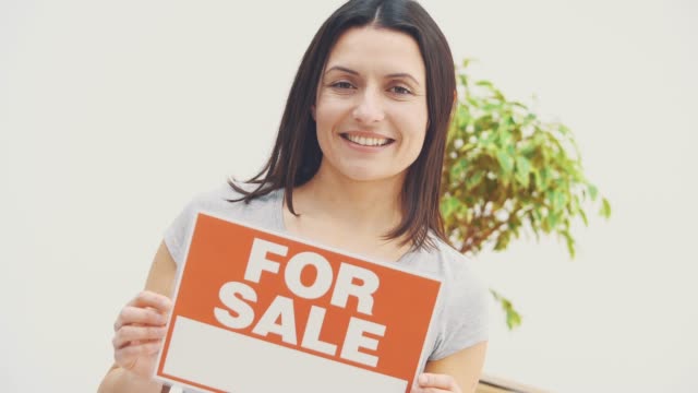 Smiling young beautiful woman is holding a plate with words FOR SALE written on it, offering house for sale and moving out.