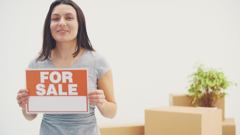 Smiling young beautiful woman is holding a plate with words FOR SALE written on it, giving thumb up.