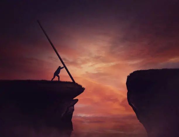 Photo of Businessman pushing a long beam, creating an improvised bridge to cross the abyss obstacle. Cover the gap and reach other side of the cliff. Mission accomplishment, overcome and success concept.