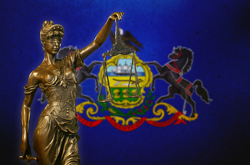 Close-up of a small bronze statuette of Lady Justice before a flag of Pennsylvania.