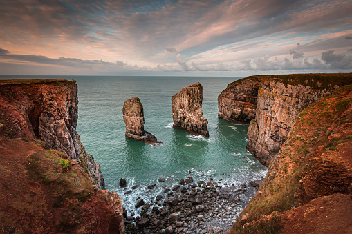 Stack rocks photographed at sunrise on dramatic coastline of Pembrokeshire ,South Wales,UK.Moody, colourful sky over bay with turquoise water, rock formation and cliffs lit by morning light.Tranquil seascape scene.