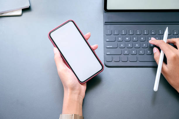 istock Top view female hand holding smartphone with blank screen and working with tablet. 1193897884