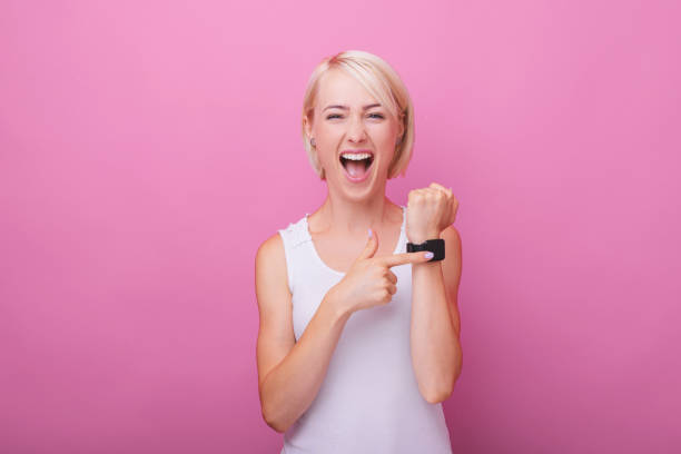 foto di donna bionda eccitata, che punta al suo orologio, in piedi su sfondo rosa isolato - checking the time women impatient wristwatch foto e immagini stock