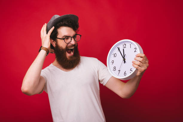 istock Young bearded hipster holding a white clock and watching it shocked, screaming and stressed on red background. 1193903443