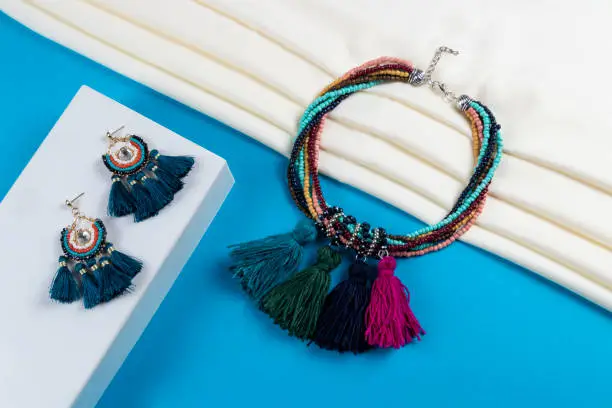Photo of set of tassel earrings and multi layer multi color chunky seed beads with colorful tassels and crystal beads necklace boho style on white scarf and blue paper background