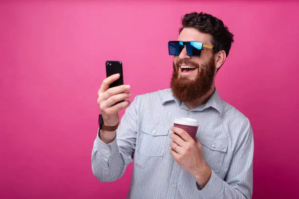 Young bearded hipster man taking a selfie with his cup of coffee on pink background.