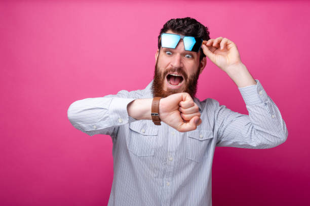 Young bearded man looking shocked at his watch, lifting up his sunglasses on pink background. stock photo