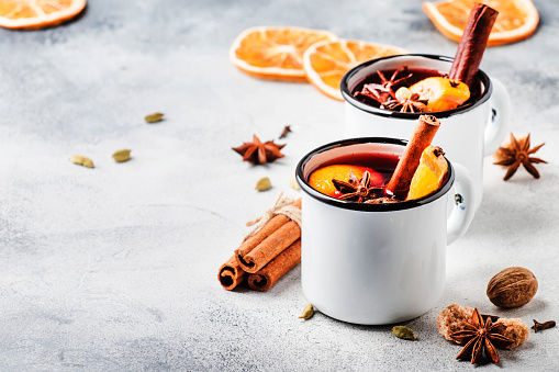 Mulled wine in white metal mugs with cinnamon, spices and orange on concrete background, traditional drink on winter holiday. Copy space for text
